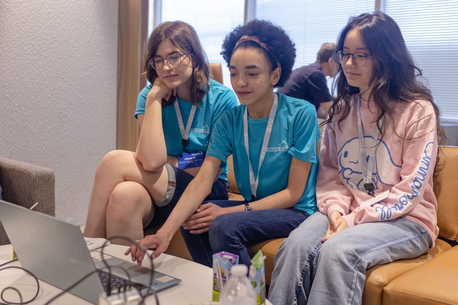 Three young women sit on a couch, focused on a laptop. Two wear matching blue shirts and lanyards, while the third wears a pink sweatshirt. The mood is collaborative.