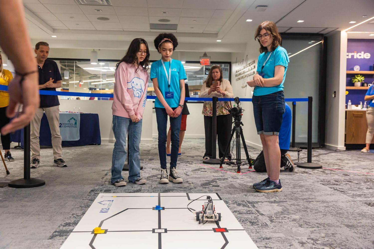 Three focused individuals watch a small robot navigate a grid on the floor. A few onlookers and a camera are in the background. It's a robotics competition.