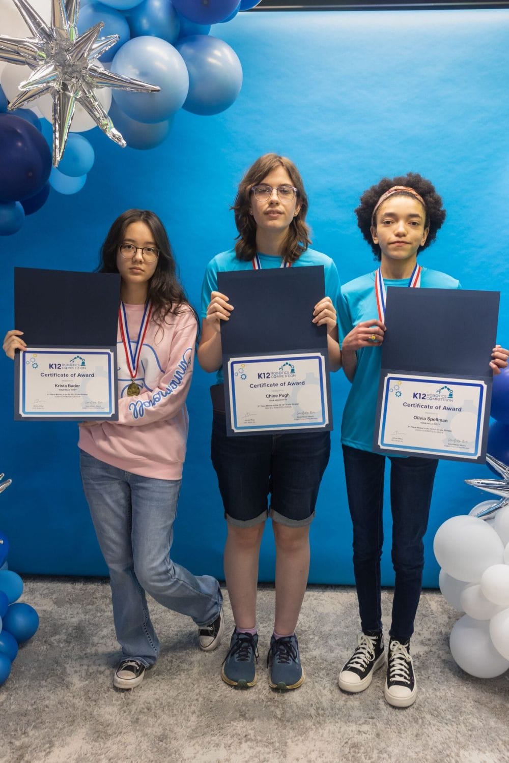 Three students stand side by side holding certificates, wearing medals. They are surrounded by blue and white balloons, exuding a celebratory mood.