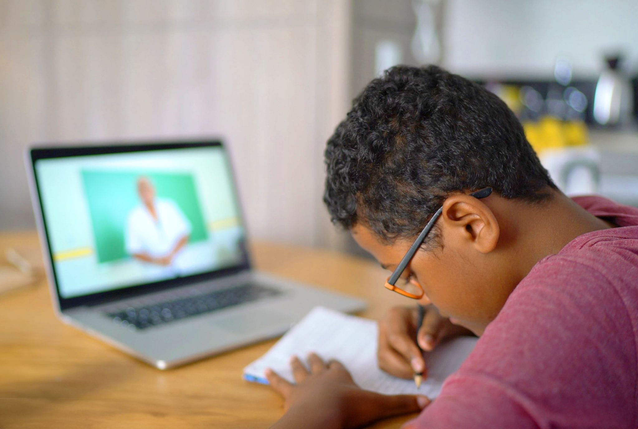 A young boy writes in a notebook while focused on a laptop screen, showcasing a moment of learning and creativity.