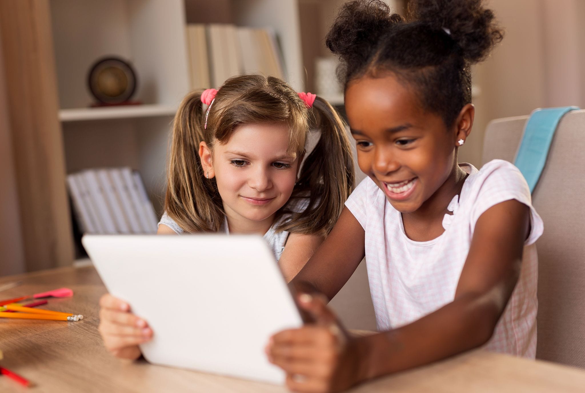 Two young girls are seated at a table, engaged with a tablet, sharing a moment of curiosity and learning together.
