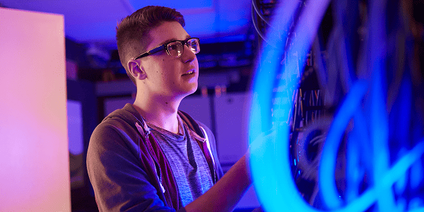 A young man wearing glasses is focused on working at his computer, typing and looking at the screen intently.