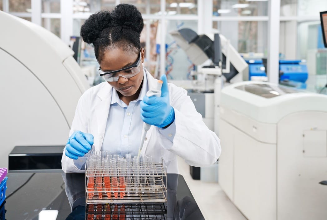 A scientist in a lab coat and safety goggles uses a pipette with test tubes.