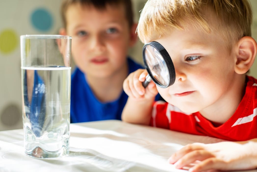 Young boy in red shirt examines a glass of water with a magnifying glass, while another boy in blue shirt watches. Curious and playful atmosphere.