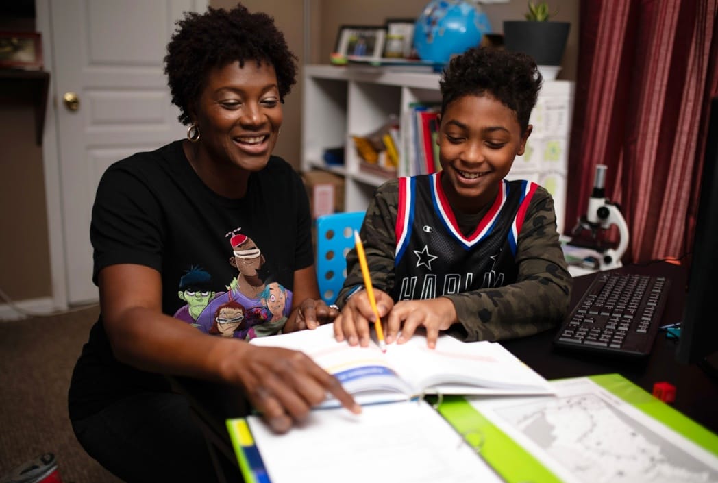 A smiling woman and boy work together on homework at a desk. The boy holds a pencil, while the woman points at a textbook, creating a cheerful, collaborative learning atmosphere.