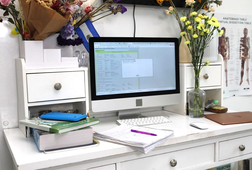 A cluttered white desk with a computer monitor displaying a data screen. Books, notes, and a pen lie nearby. Colorful flowers are in vases, adding a lively touch.