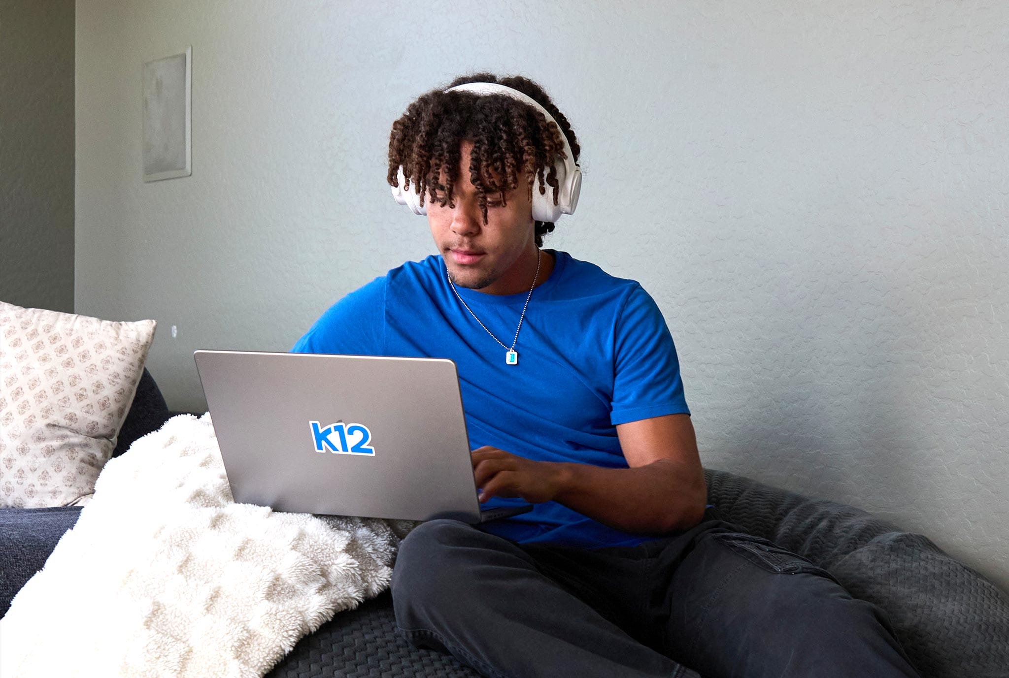 Young man in a blue shirt wears headphones, focused on a laptop with "k12" on it, sitting on a cozy couch with pillows. Calm, studious atmosphere.