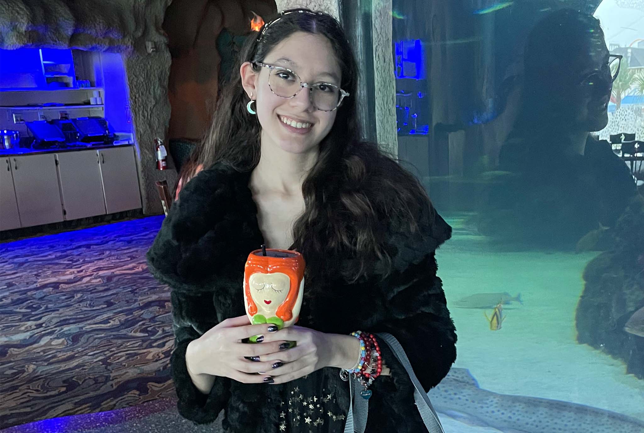 Young woman in a black fur coat holds a colorful ceramic mug with a serene expression. She stands in a dimly lit room, smiling warmly.