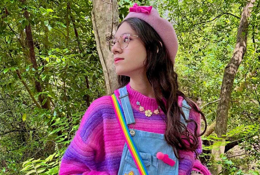 Young woman in a vibrant pink and purple sweater with overalls, wearing glasses and a pink beret. She stands amidst lush green trees, exuding a playful vibe.