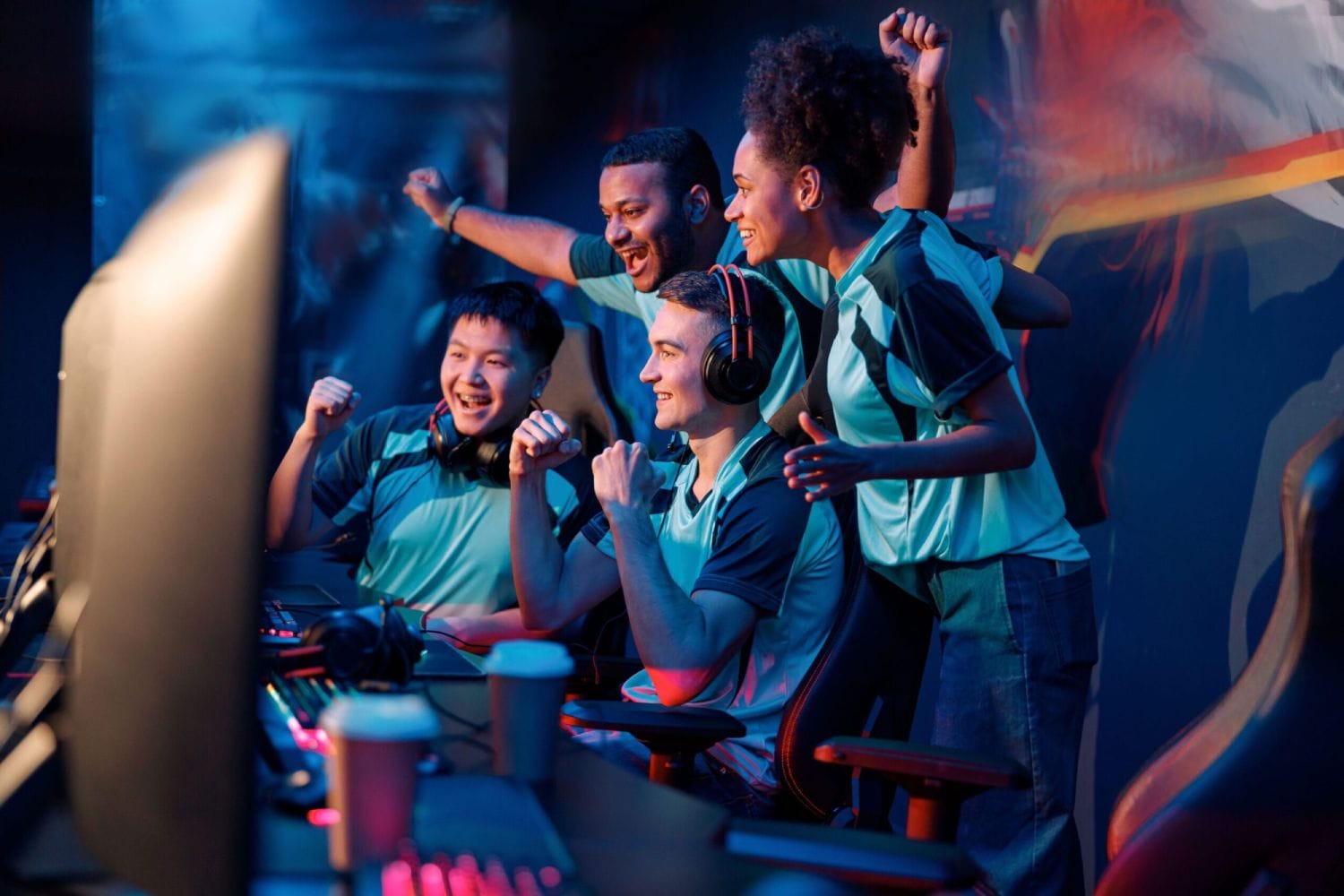 A diverse group of four esports players wearing matching jerseys celebrate triumphantly in front of computer screens, conveying excitement and teamwork.