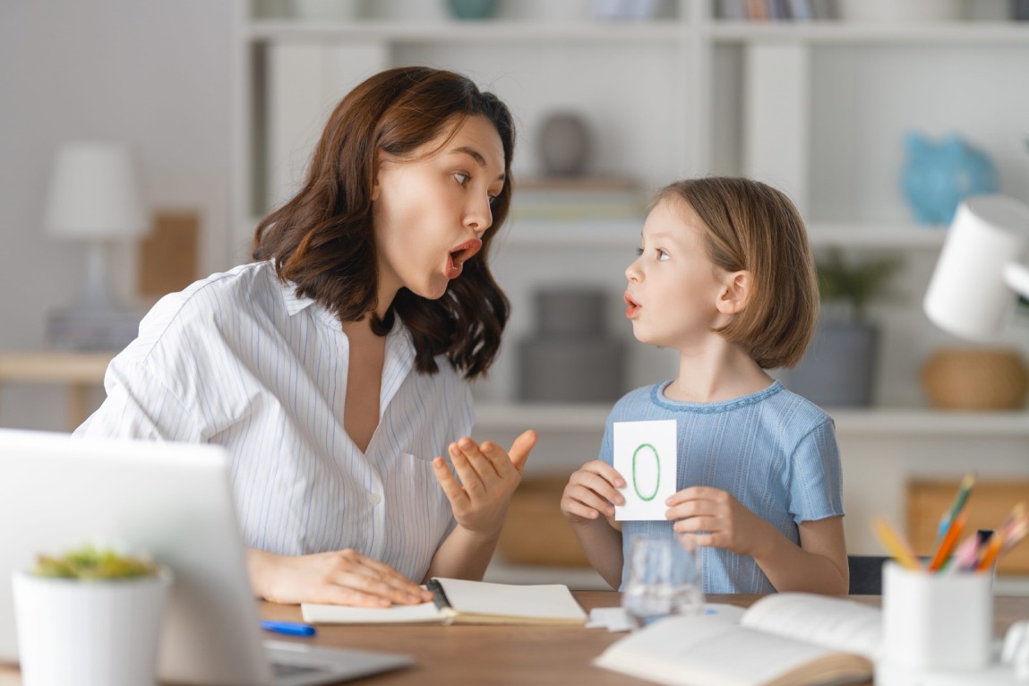 A woman and a child engage in a learning activity at home. The child holds a card displaying the letter "O." Both are animated and expressive.