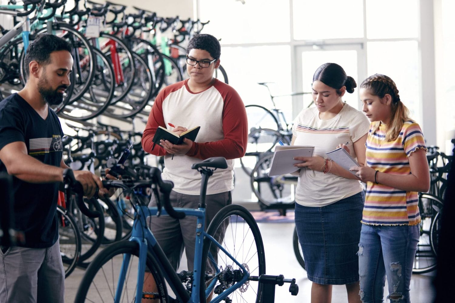 Man demonstrating bike assembly to three attentive students taking notes in a well-lit bike shop. Background features rows of colorful bikes.