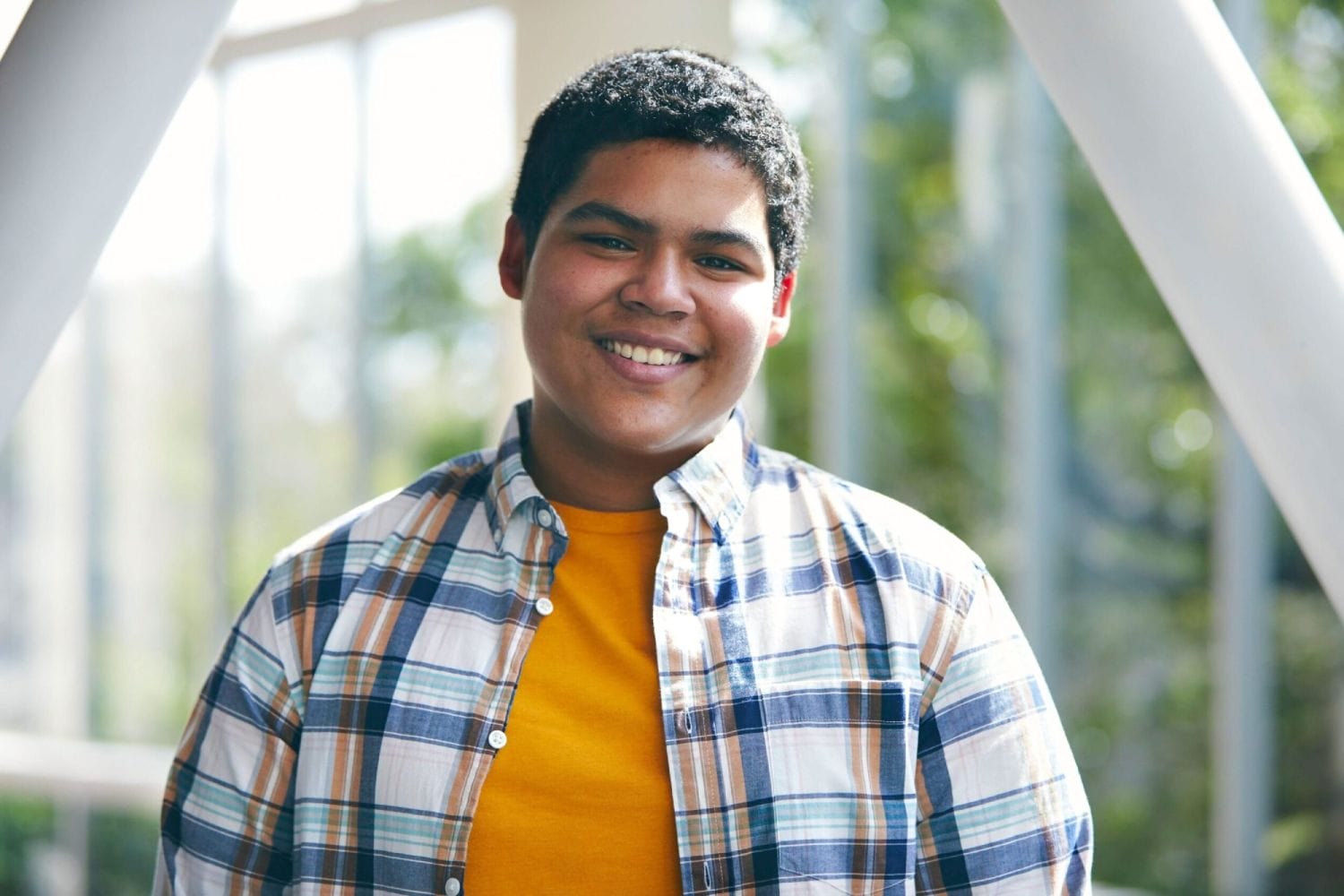 Smiling person in a plaid shirt and yellow T-shirt stands indoors with sunlight filtering through large windows, creating a warm, inviting atmosphere.