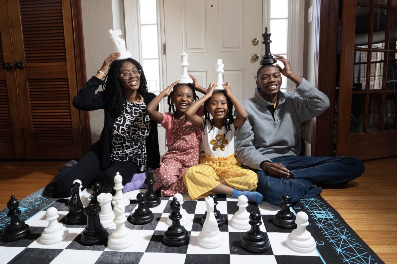 Smiling family seated on floor, each holding a large chess piece on their head. A chessboard with black and white pieces is in front of them, creating a playful atmosphere.