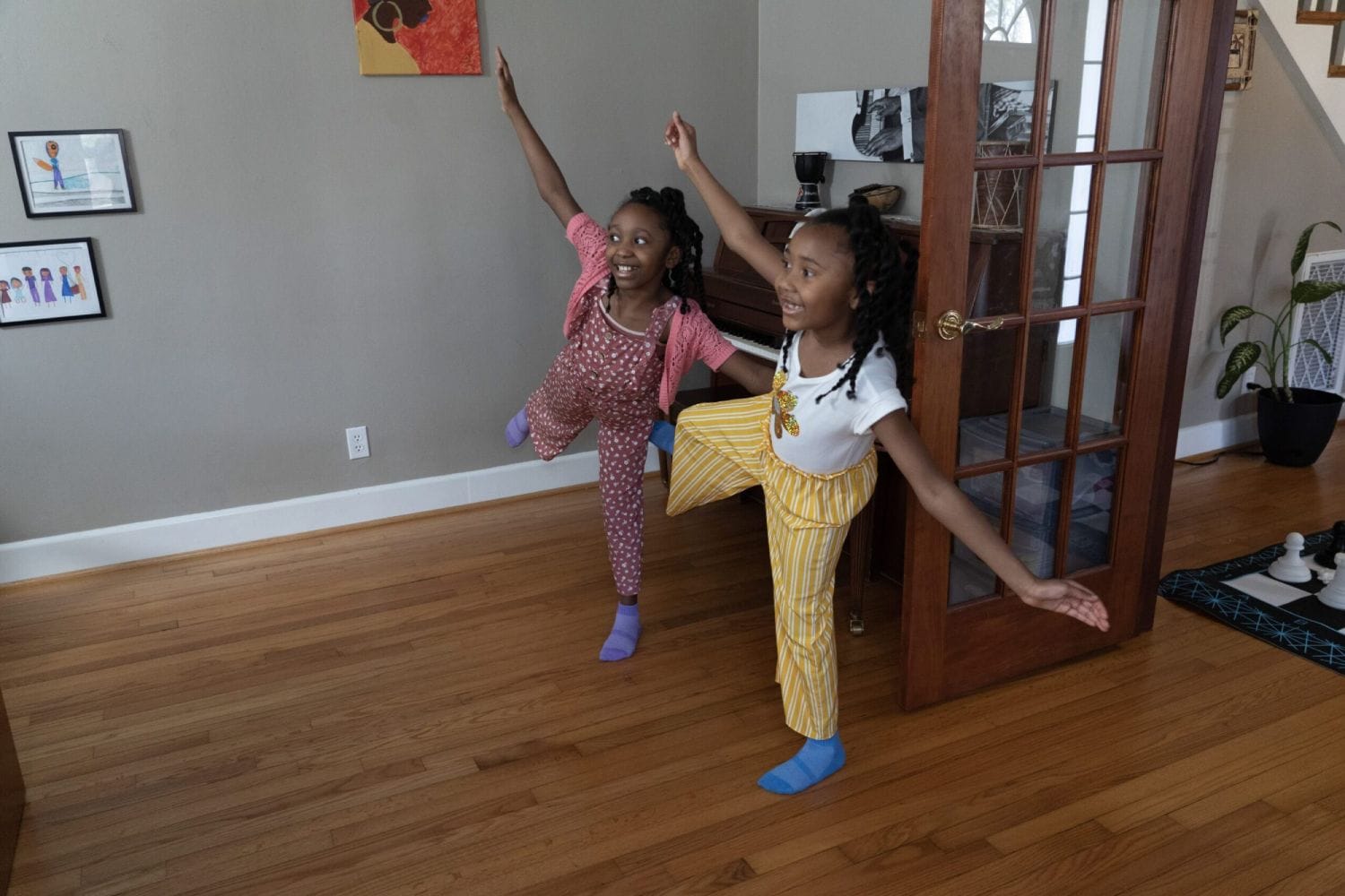 Two smiling children strike playful poses on a wooden floor in a living room. Framed art decorates the walls, adding a cheerful atmosphere.