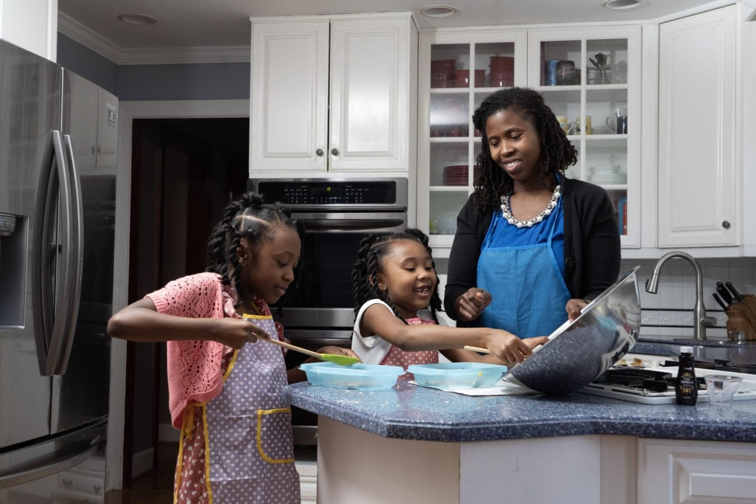A woman and two children happily cook together in a bright kitchen. The children wear aprons and eagerly mix ingredients, creating a joyful atmosphere.