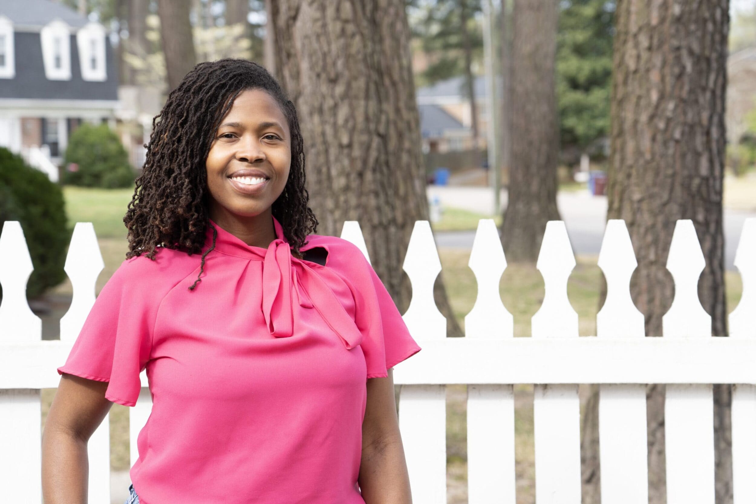Smiling woman with curly hair wearing a pink blouse stands in front of a white picket fence. Background shows trees and a house, conveying a cheerful and relaxed atmosphere.