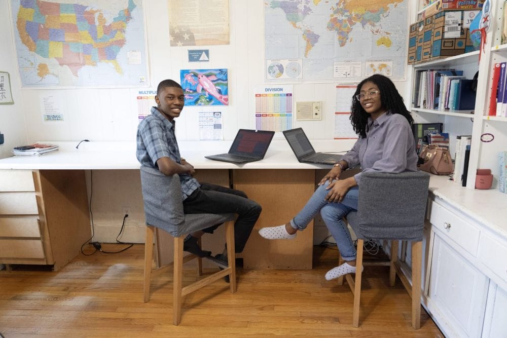 Two people sit on chairs at a desk with laptops in a study area. Maps and educational posters decorate the walls. They appear relaxed and engaged.
