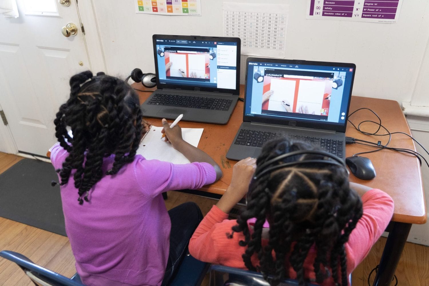 Two children with braided hair engage in an online art class using laptops. One writes on a notebook while the other wears headphones, focusing intently.