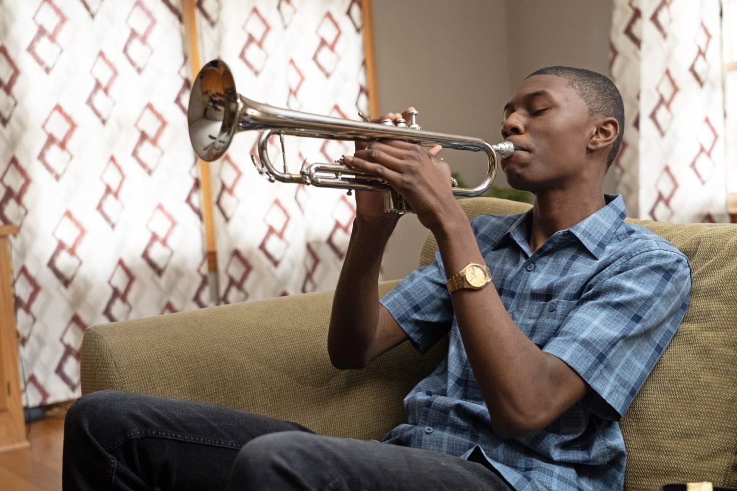 A young man plays a trumpet on a couch, wearing a blue checkered shirt and a gold watch. The room has patterned curtains, creating a calm atmosphere.