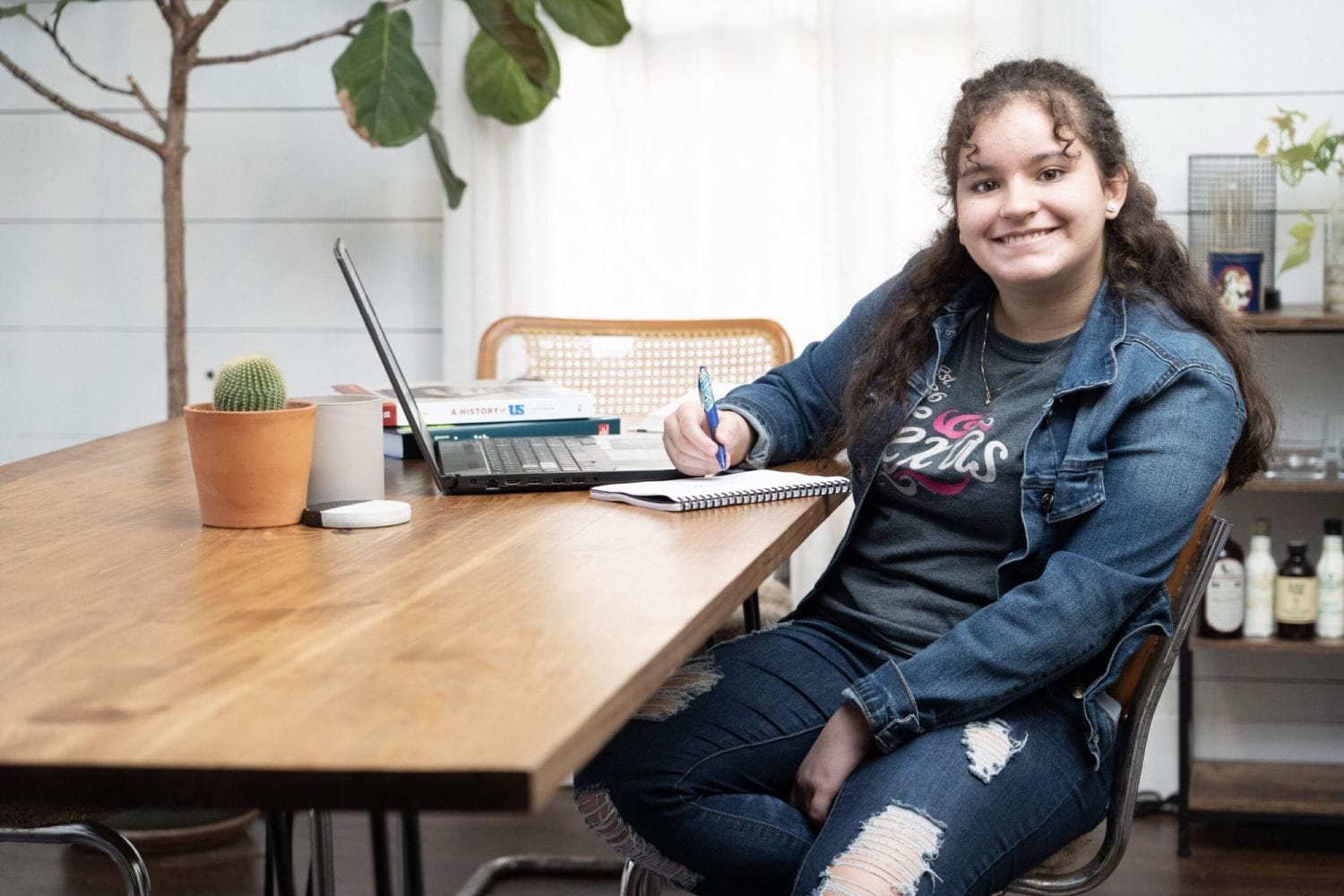 Young woman smiling, sitting at a wooden table with a laptop, books, and a notebook. She wears a denim outfit, conveying a relaxed study environment.