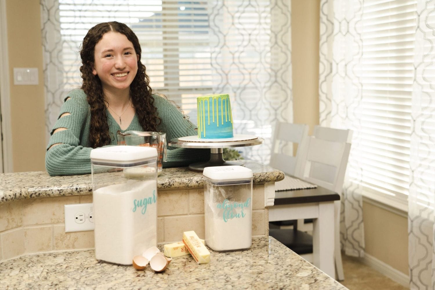 A young woman smiles in a kitchen, next to a blue and green drip cake on a stand. Nearby are containers of sugar and almond flour, with eggshells and butter.