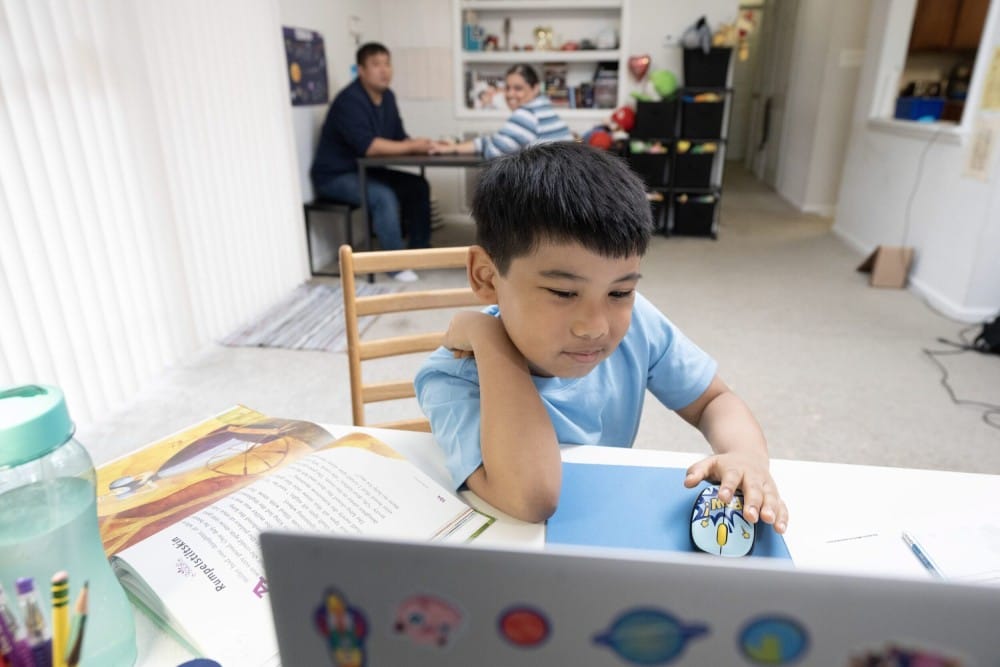 Young boy in a blue shirt uses a mouse at a laptop, focused on the screen. Open book and water bottle nearby. Two adults converse in the background, adding a warm, supportive tone.