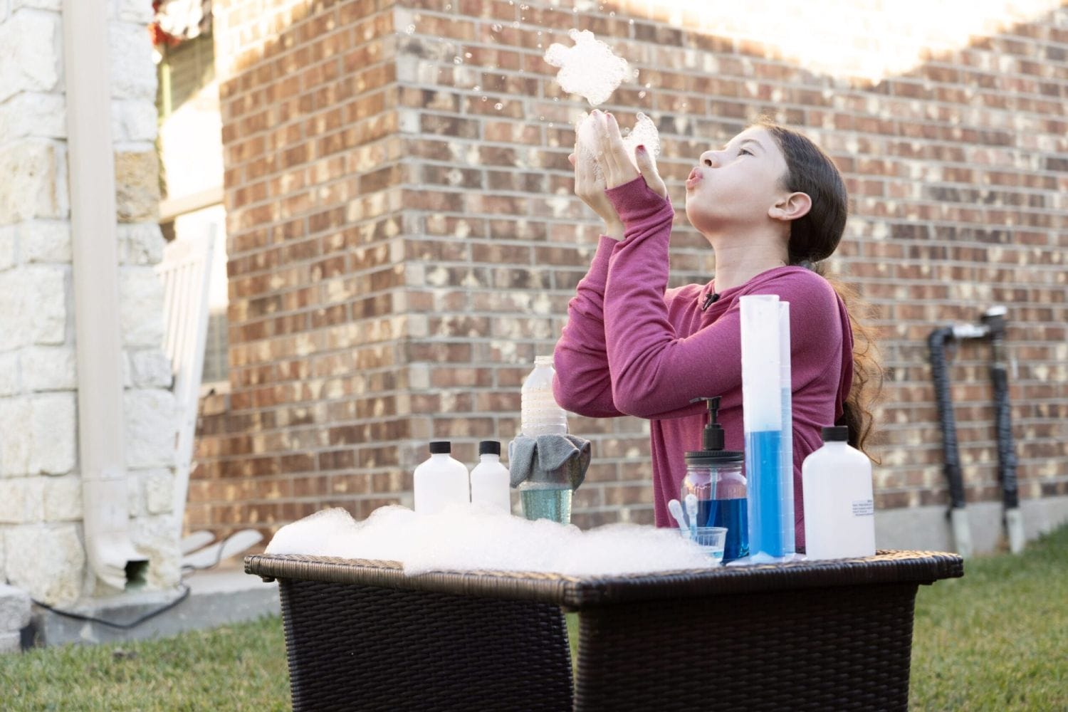 A young girl in a pink sweater joyfully blows bubbles outdoors. She is seated near a wicker table with bubble-making supplies and bottles, set against a brick wall background.