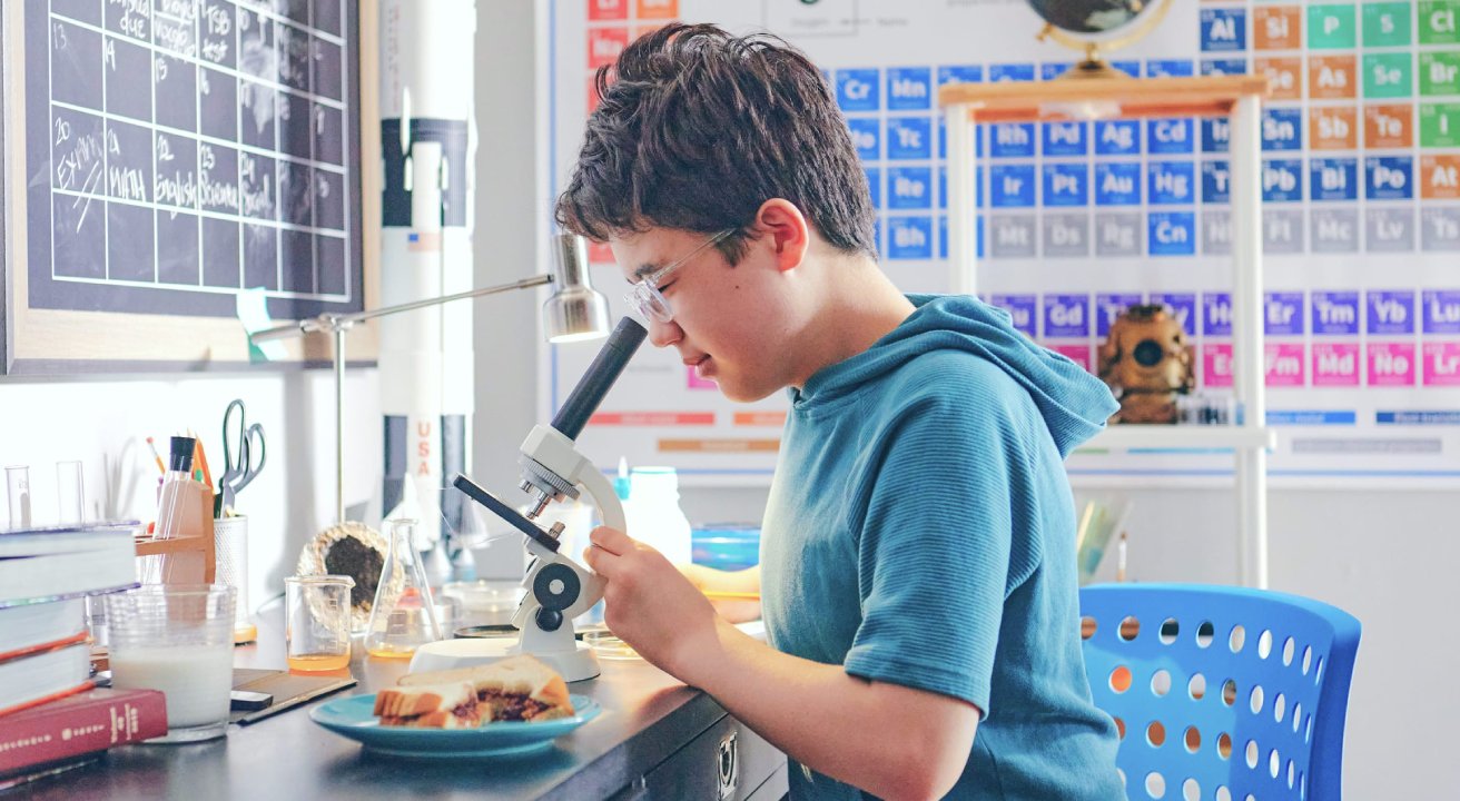 Young person in blue hoodie uses a microscope at a science-themed desk with beakers. A periodic table and blackboard are visible in the background.