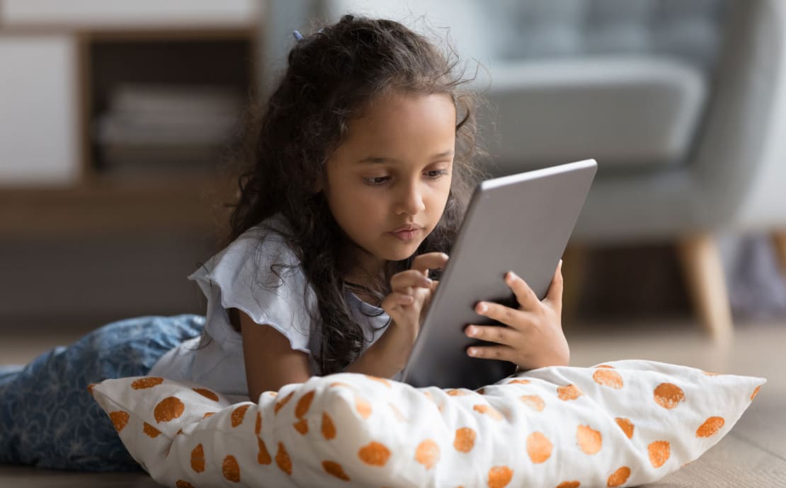 A young girl with curly hair intently focuses on a tablet while lying on a pillow with orange dots. She is in a cozy living room setting.