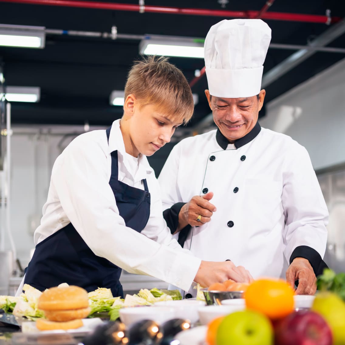 Two chefs in a bright kitchen prepare food. One, in a black apron, focuses intently, while the other, wearing a tall hat, smiles supportively.