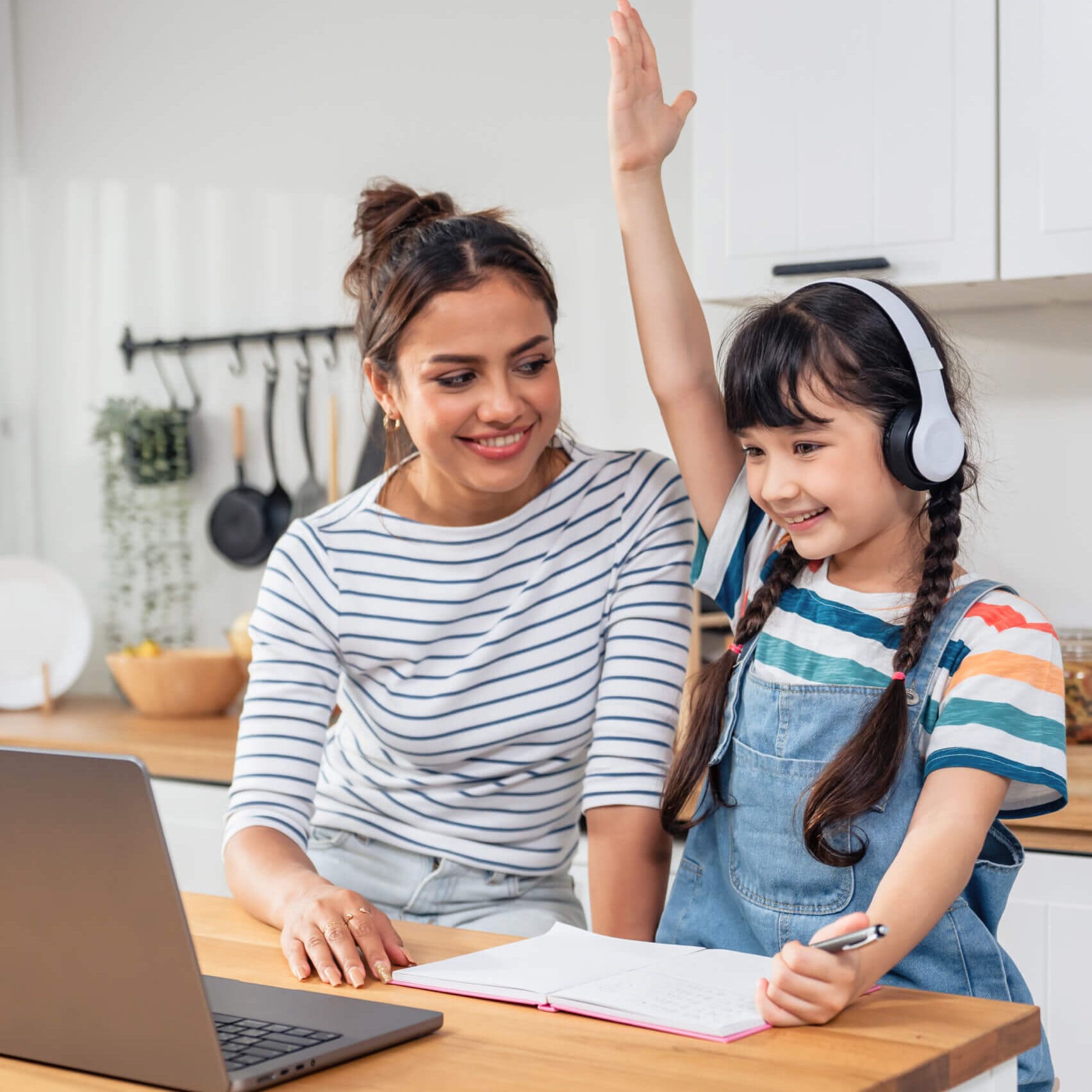 A smiling woman in a striped shirt sits beside a young girl with headphones and raised hand, working at a table with a laptop and notebook in a kitchen.