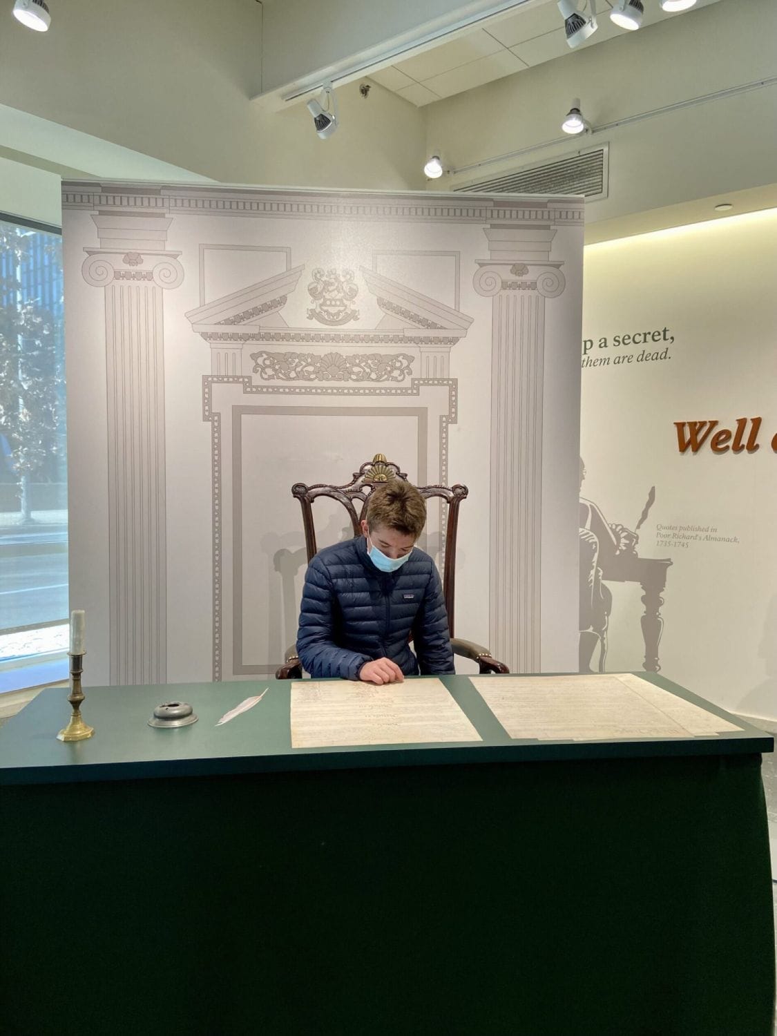 A person in a blue jacket and mask sits at a green desk, reading large documents. The background features classical columns and a quote on the wall.