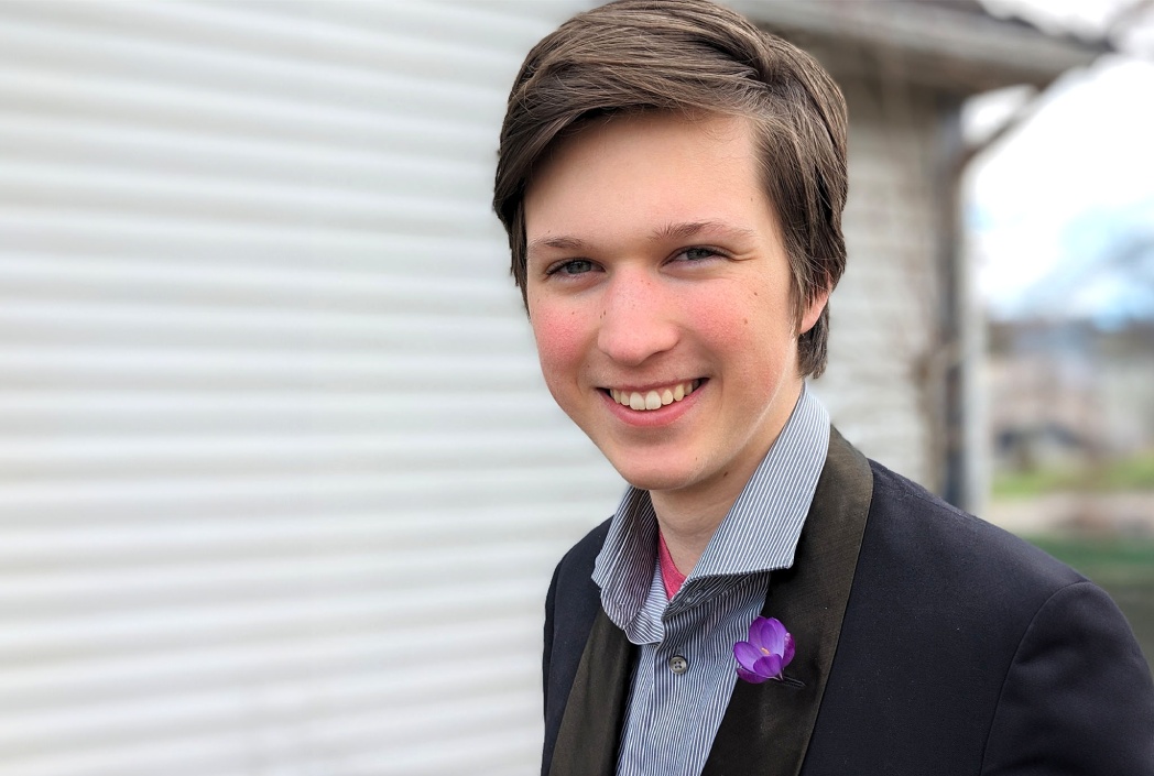 Smiling person outdoors, wearing a dark jacket with a purple flower pin. Background shows blurred siding and greenery, conveying a casual, cheerful mood.