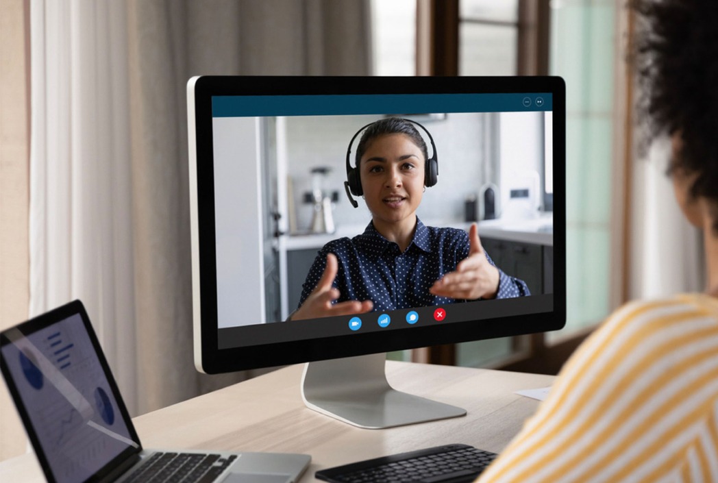 A person in a striped shirt participates in a video call. The monitor shows a person wearing headphones, gesturing while speaking in a well-lit room.