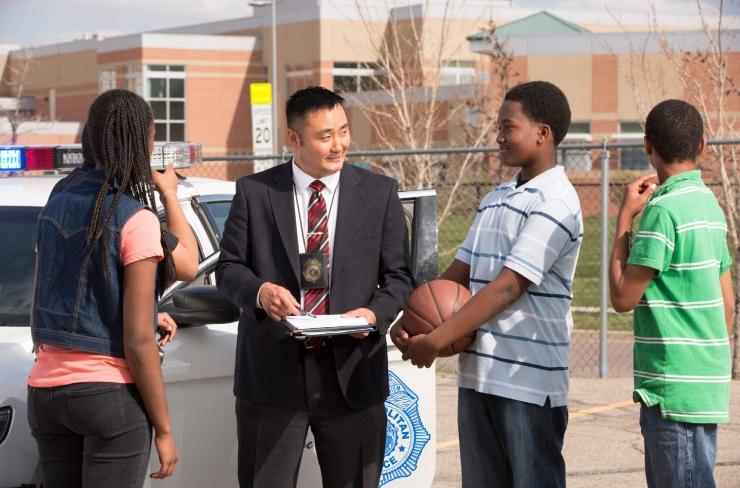 A police officer talks to three teenagers near a patrol car. One teen holds a basketball. The scene appears collaborative and positive.