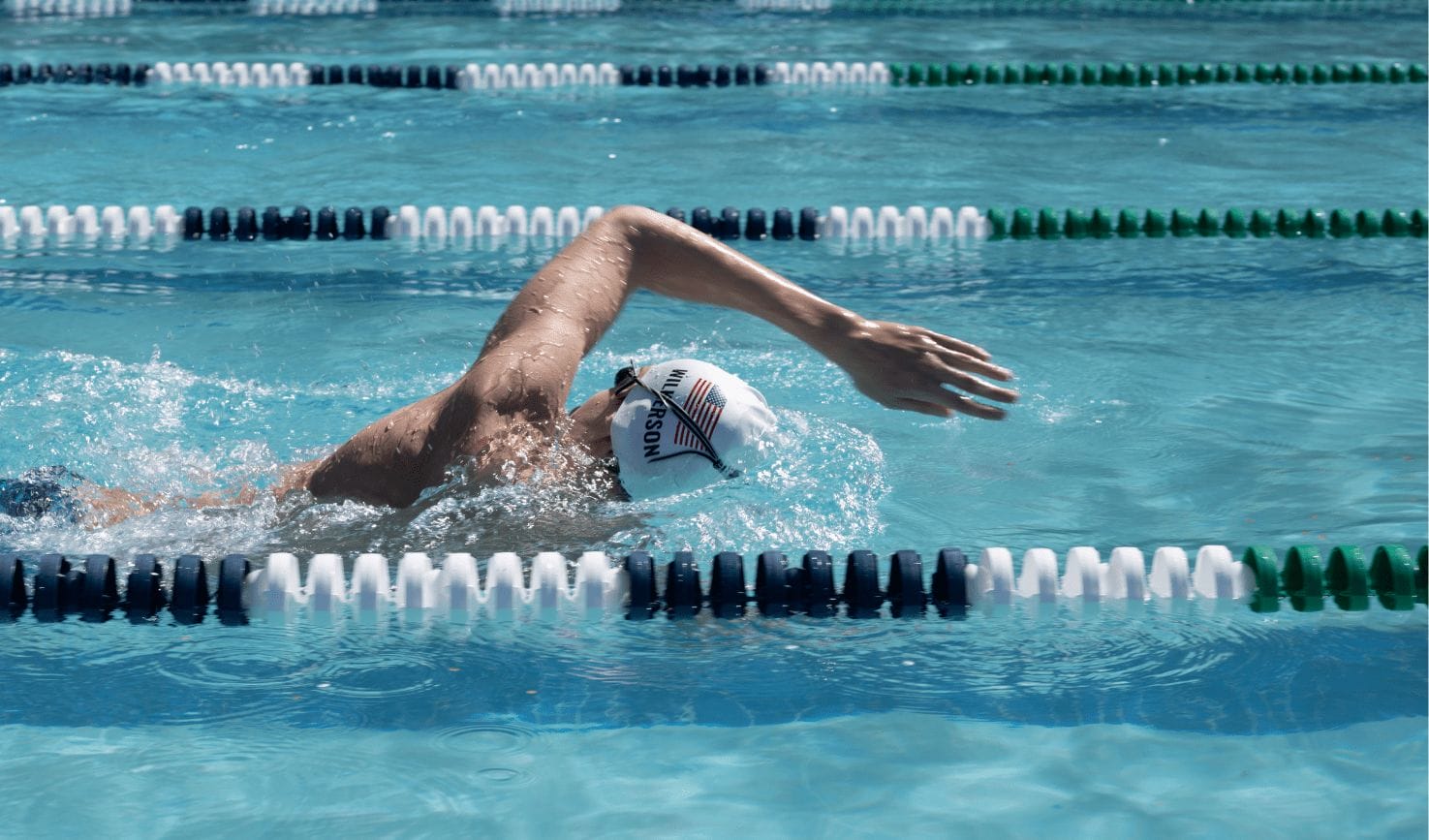 A swimmer in a white cap performs a freestyle stroke in a clear blue pool. White and green lane dividers are visible, indicating a competitive setting.