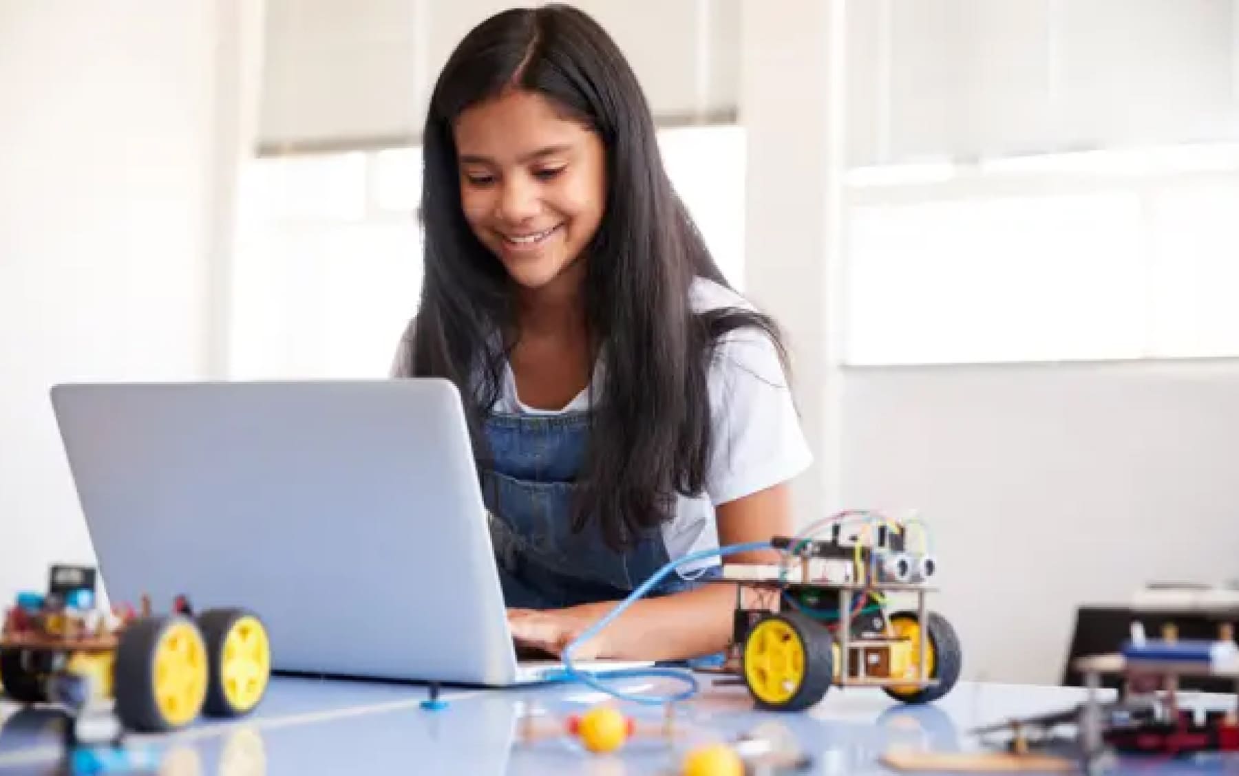 A young girl smiles while working on a laptop at a table with robotics projects. Bright, sunny room conveys a sense of creativity and learning.