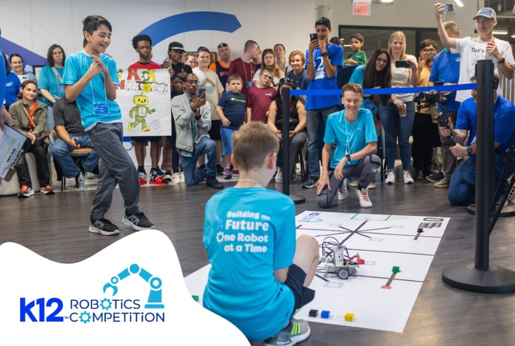 A young boy kneels on a robotics competition mat, excitedly operating a robot. A crowd of spectators watches, capturing the moment with cameras.