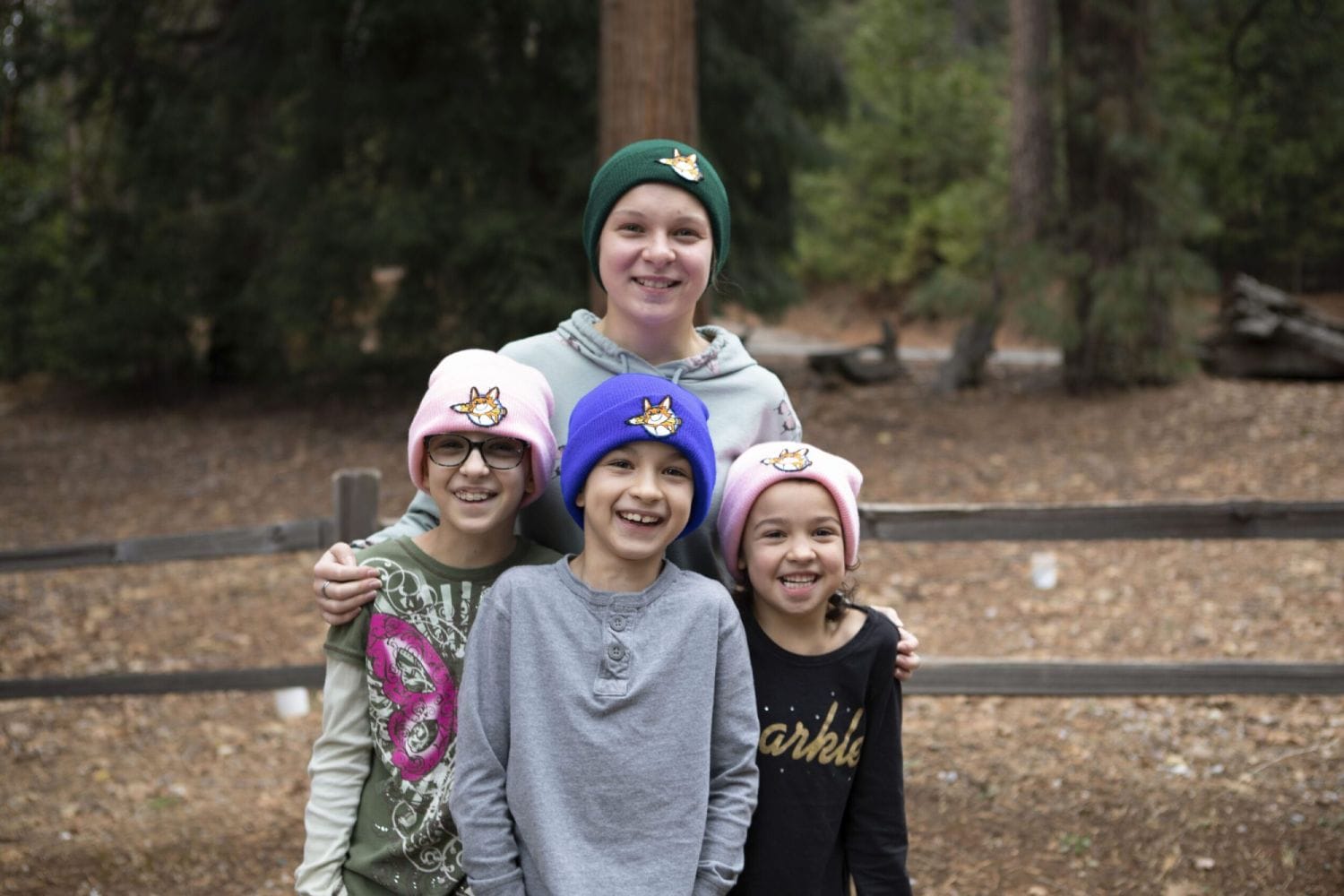A smiling group of four children wearing colorful beanies with animal patches. They're standing in a forest, radiating warmth and joy.