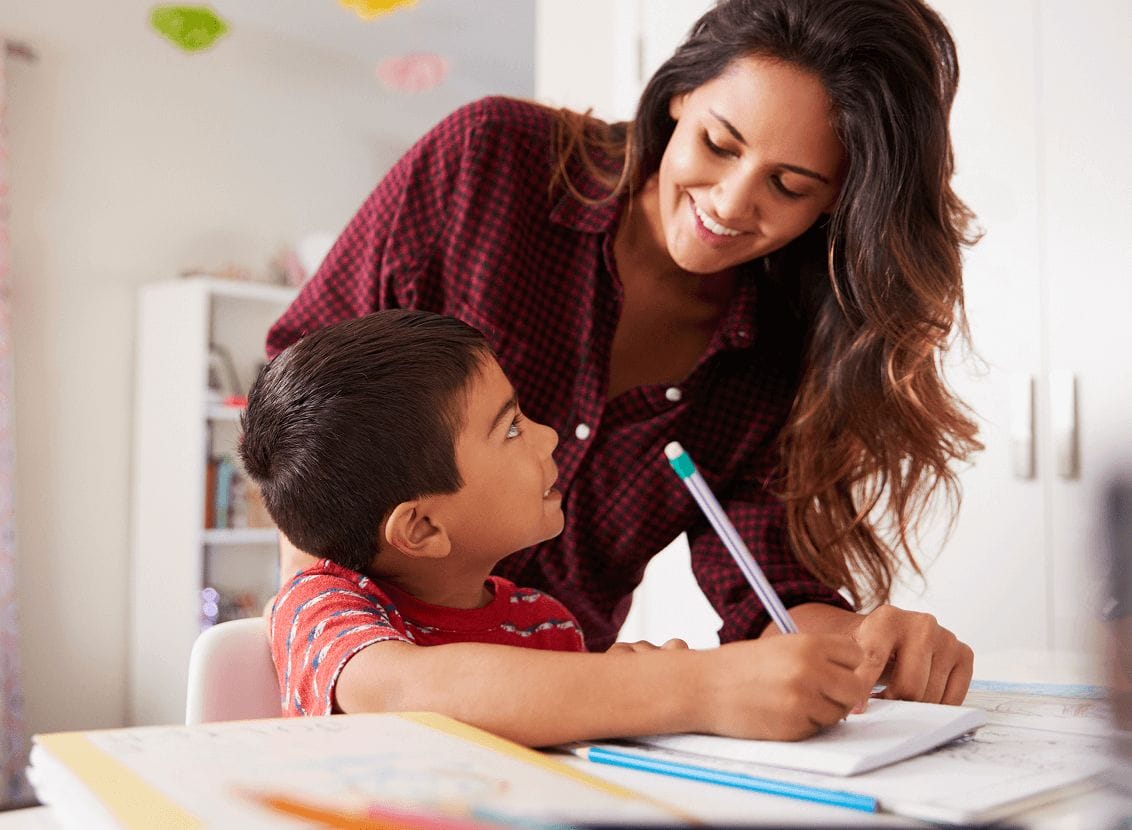 A woman in a checkered shirt smiles at a young boy as she assists him with homework. The boy writes in a notebook, looking up at her warmly.