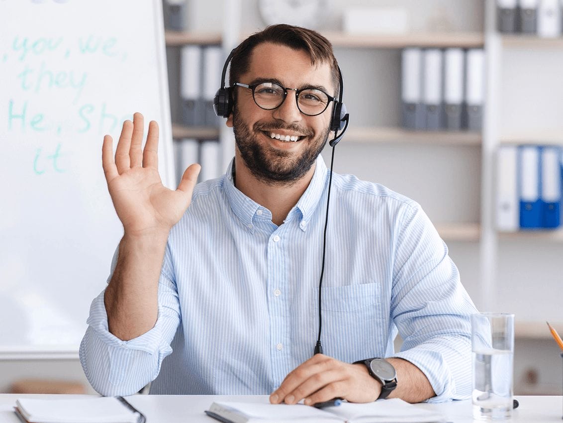 Smiling man wearing glasses and headphones waves at his desk with notebooks and a glass of water. A whiteboard with words is blurred in the background.