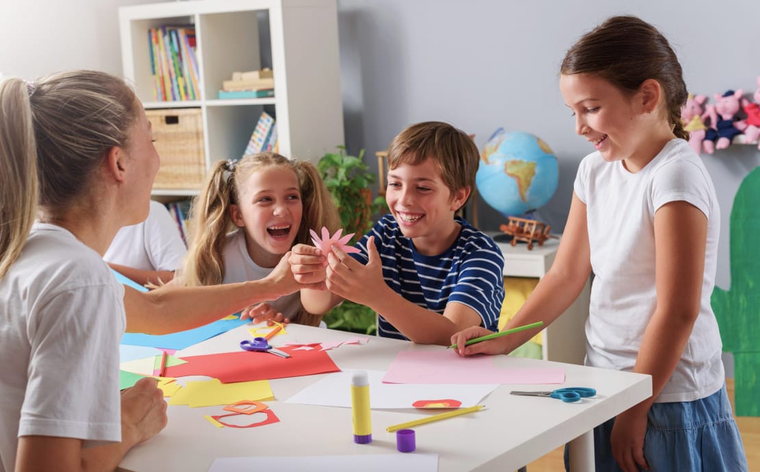 Children and a teacher are joyfully working on a craft project at a table. The room is bright and colorful, with art supplies, a globe, and toys in the background.