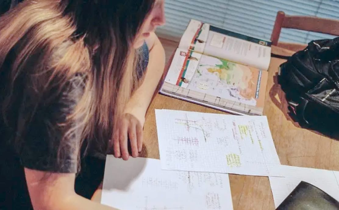 A person with long hair studies at a table with open textbooks, graph paper with notes and diagrams, and a black backpack. The scene conveys focus and concentration.