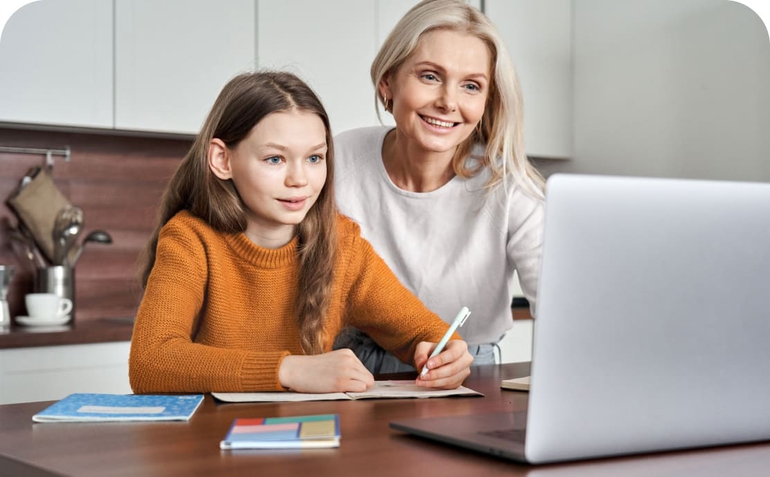 A smiling woman and a girl in a cozy kitchen work together on homework. The girl writes, using a laptop, while colorful notebooks lie on the table.