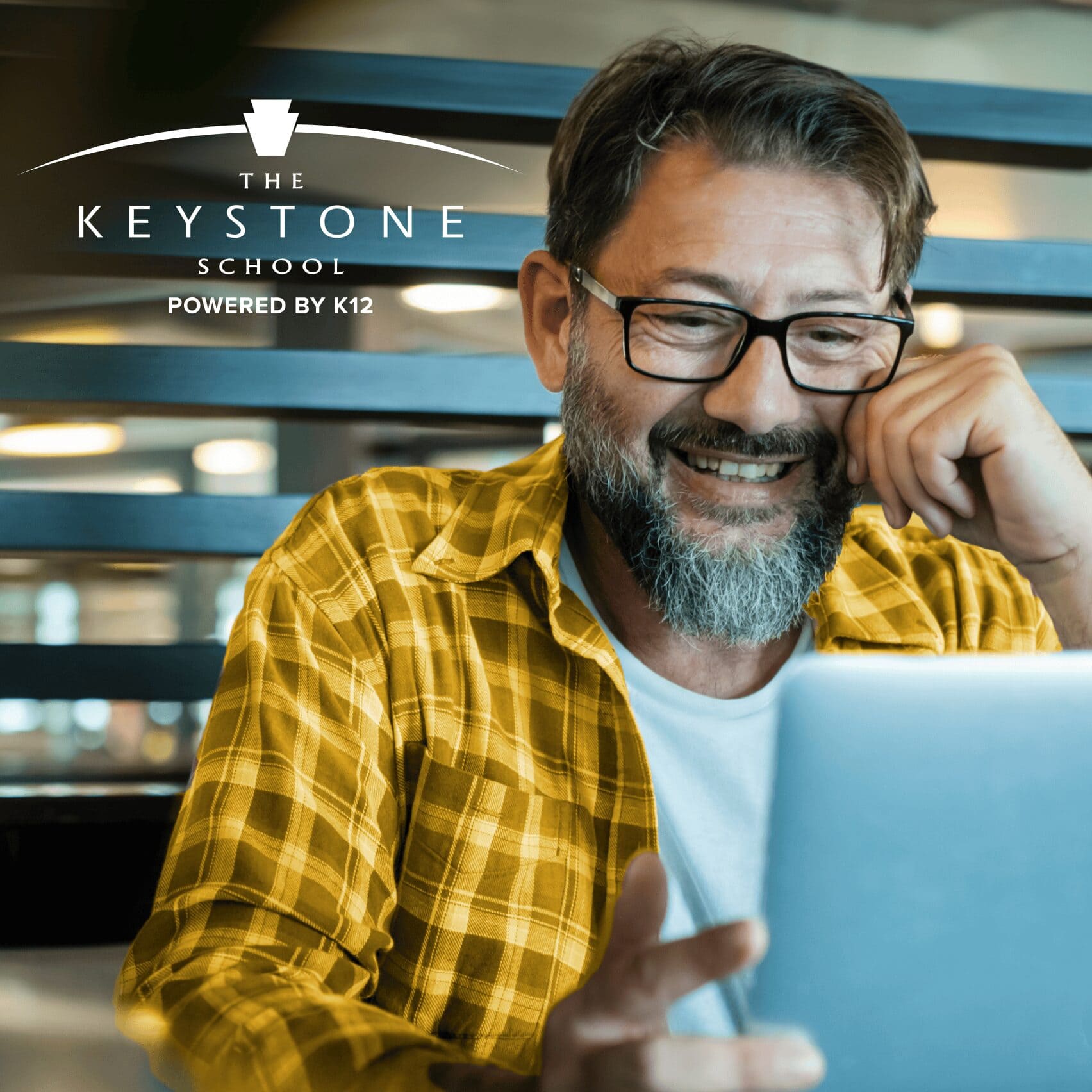 A man with glasses and a plaid yellow shirt smiles at a laptop. The Keystone School logo is in the corner. The setting is casual and bright.