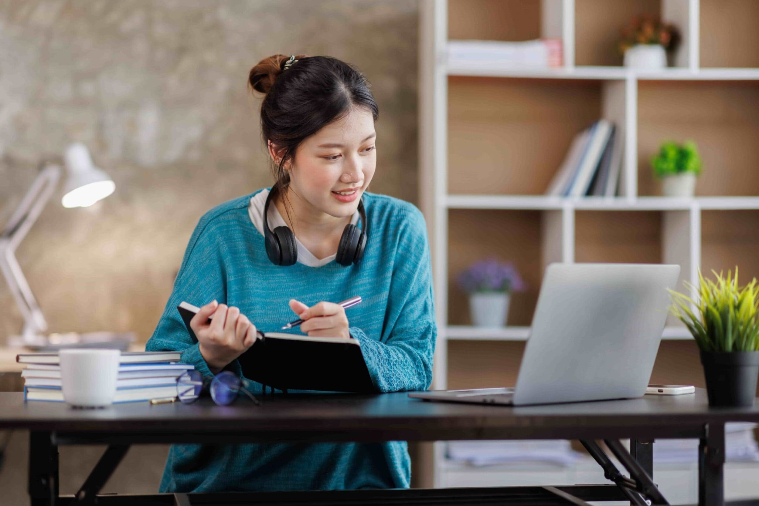 Young woman in a blue sweater sits at a desk with a laptop, notepad, and stacks of books. Headphones rest around her neck. She appears focused and engaged.