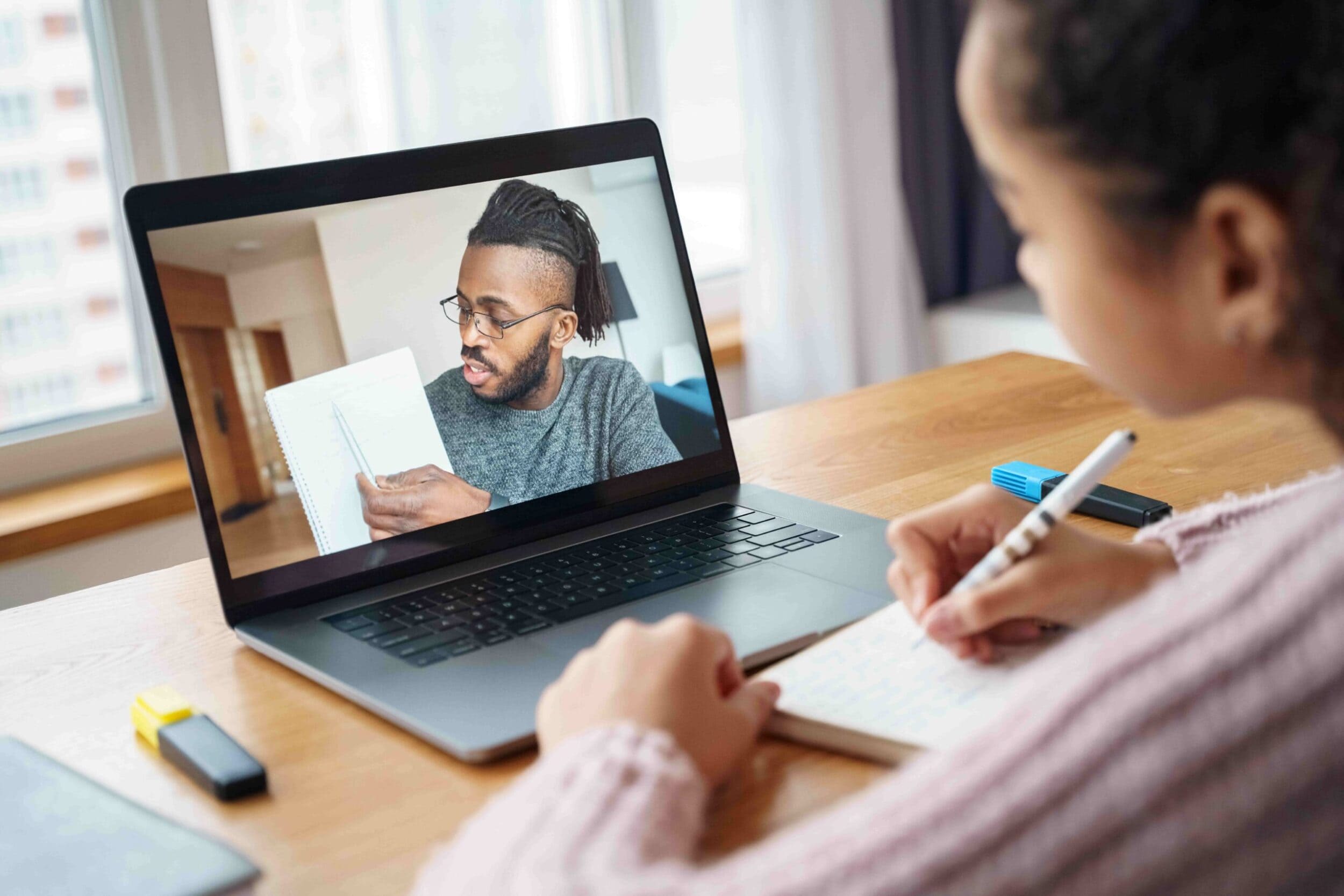 A young woman takes notes while attending an online class on a laptop. On the screen, a man holds up a notebook, explaining a concept. The scene is focused and educational.