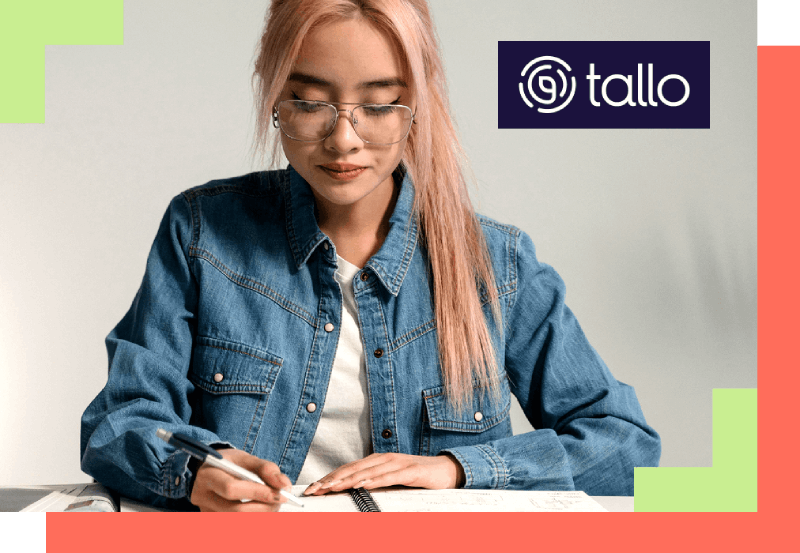 Young woman with glasses and pink hair, focused on writing in a notebook. She wears a denim jacket; "tallo" logo is in the top right corner.