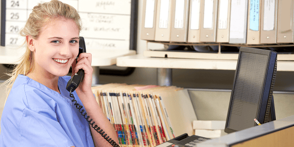 Smiling woman in blue scrubs talks on the phone at a desk, with shelves of files and a computer in a bright office setting, conveying professionalism.